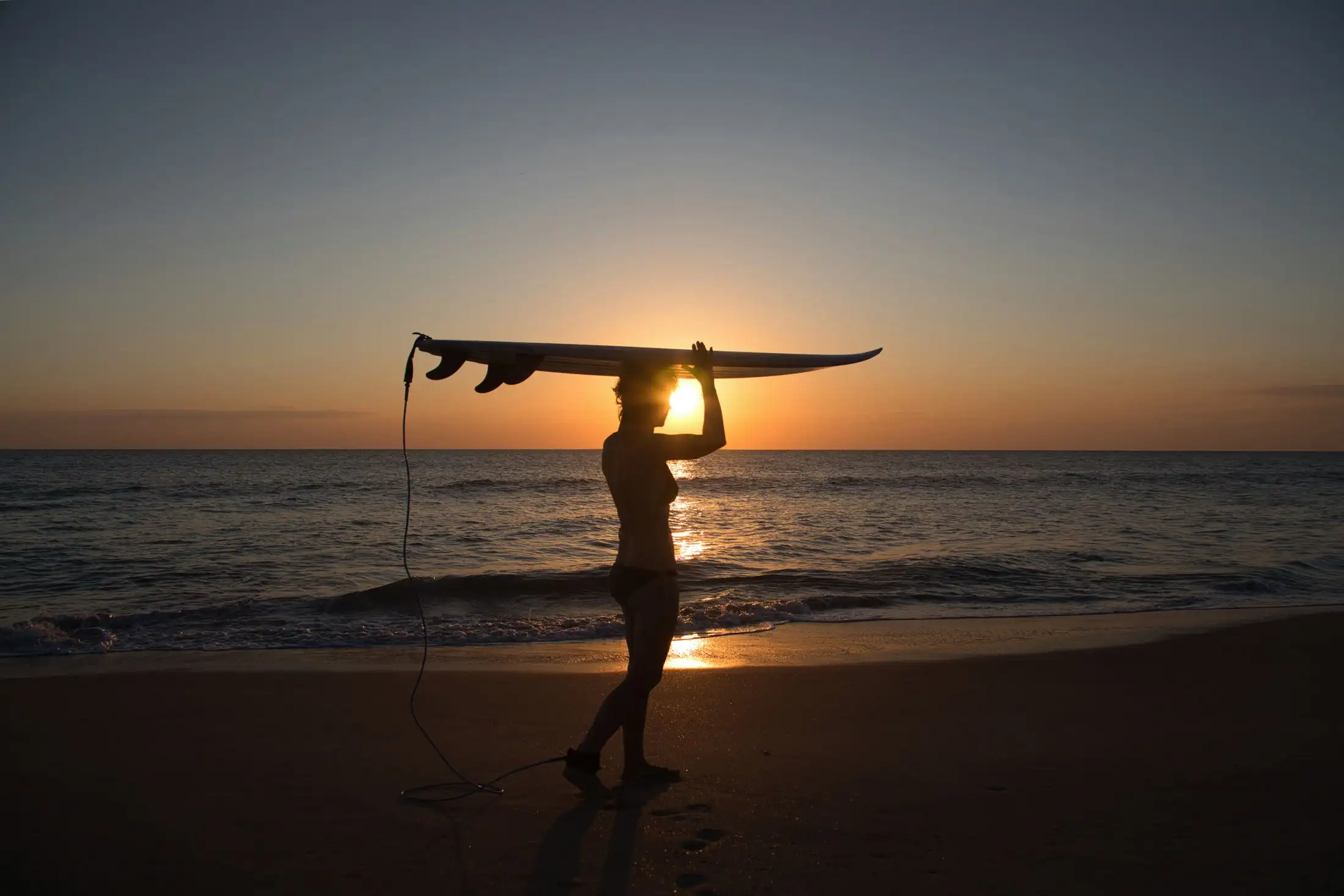 Silhueta de surfista erguendo a prancha acima da cabeça, com o sol se pondo atrás
