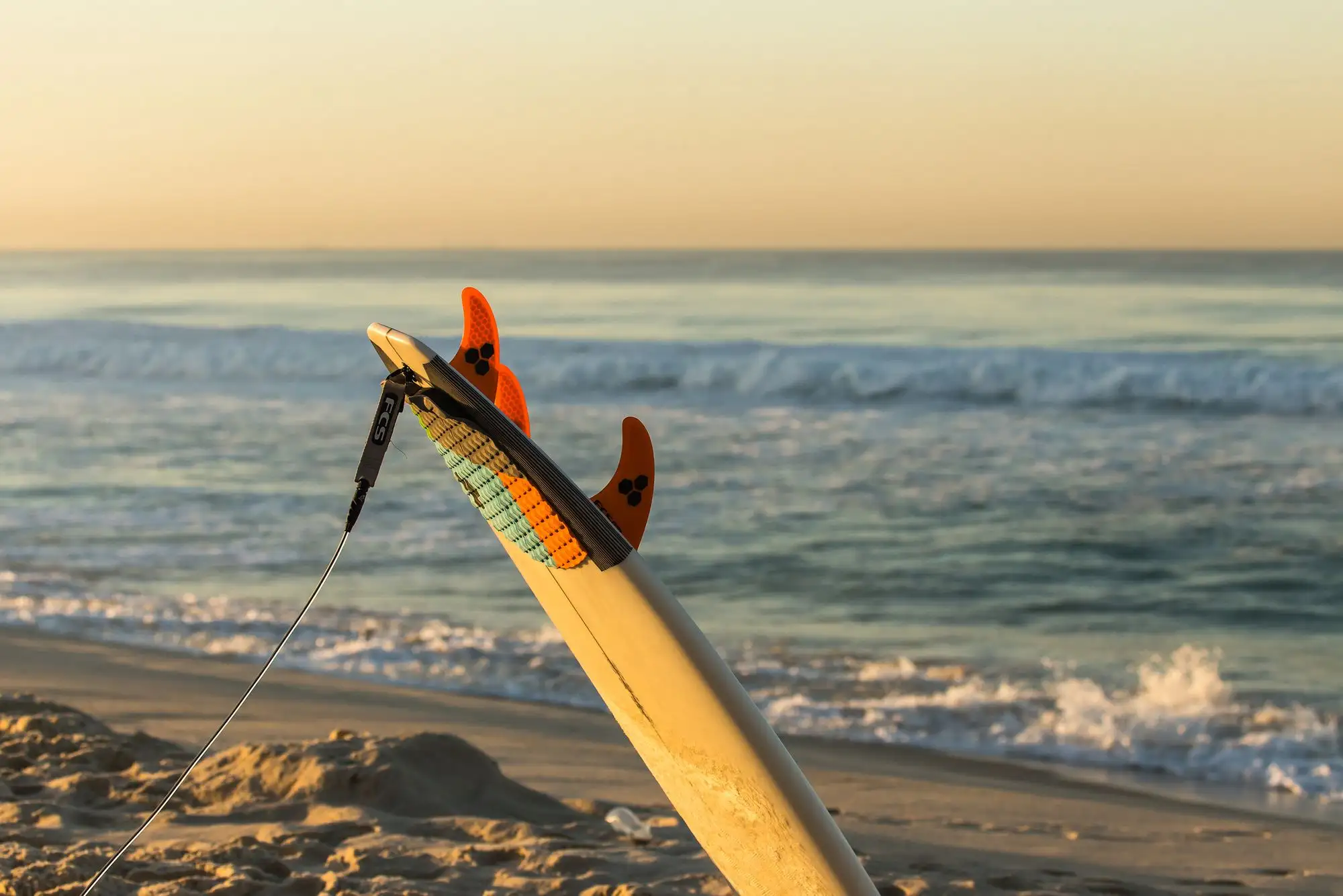 Prancha de surf fincada na areia, com as quilhas à mostra e o mar ao fundo